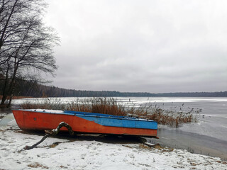 An old boat on the shore of a frozen pond - lake against the background of a forest in the snow. Ice on the lake, ice rink and winter fishing