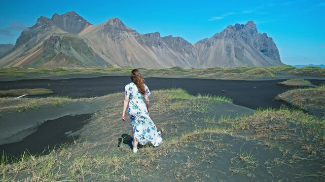 A young woman in a dress enjoys Stokksnes, at Vestrahorn Mountain, Iceland. Tourists visit tall scree mountains made of granophyre rocks. Headland with a dramatic mountainscape and black sands.