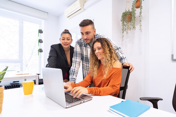 A multi-ethnic team of three collaborates around a laptop in a bright office adorned with green plants