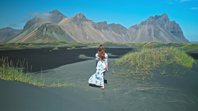 A young woman in a dress enjoys Stokksnes, at Vestrahorn Mountain, Iceland. Tourists visit tall scree mountains made of granophyre rocks. Headland with a dramatic mountainscape and black sands.