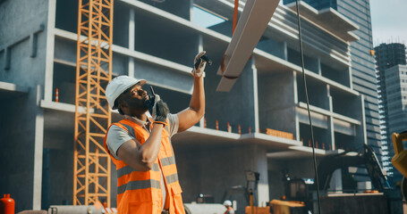Fototapeta premium Competent African American Contractor, Site Manager, Civil Engineer in a Hard Hat and Safety Vest. Black Man Helping a Crane Operator to Safely Move a Steel Beam at a Construction Site