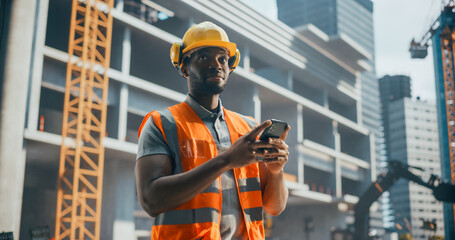 Portrait of Handsome African Builder, Worker, Craftsman Wearing a Hard Hat and a Vest while Standing at a Commercial Building Construction Site and Using a Smartphone to Browse Through Social Media