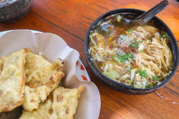 Soto Ayam and beef soto in coconut shell bowls on wooden table with mendoan snacks, satay and drinks. Traditional Indonesian soup mainly consists of broth, meat or chicken, and vegetables.