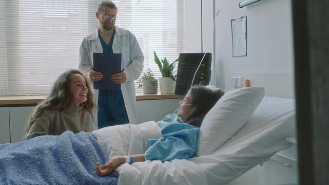 Full medium shot of male baby doctor letting Caucasian mother speak with recovering girl in hospital ward
