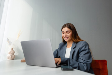 Woman in a gray blazer is looking at her laptop and smiling during an online conference. The workplace is well lit and a vase of dried plants adds aesthetic appeal. Copy space. 