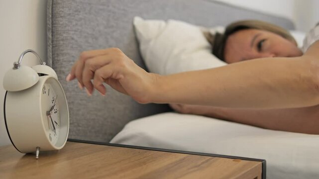 Woman wakes up in morning and turns off alarm clock on her nightstand Morning light fills room as she reaches for alarm clock Perfect scene capturing daily morning routine with focus on alarm clock