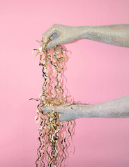 Hands adorned with glitter hold shiny streamers against a pink background.