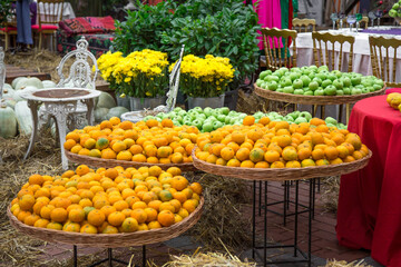 Fresh oranges and green apples are neatly arranged in wicker baskets at an outdoor market. Vibrant yellow flowers and rustic decor create a cozy atmosphere.