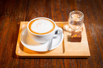 Wooden tray with copy space featuring a cup of cappuccino with whipped milk cap, a glass of water, a spoon and a piece of brownie