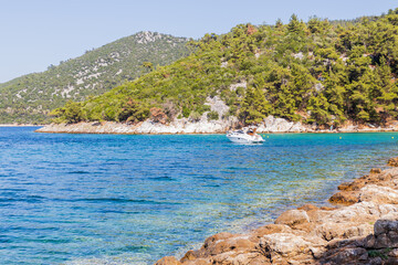 Obraz premium Sleek white yacht anchored in crystal-clear waters, near a rocky, tree-lined coastline under a sunny sky.