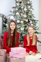 Two Girls Sitting by Christmas Tree with Gifts. Two young girls in matching red dresses sitting near a beautifully decorated Christmas tree, surrounded by pink and gold gift boxes.