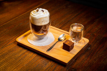 High angle shot of a glass of coffee topped with whipped cream and coffee beans, a glass of water, spoon and a brownie on a wooden tray