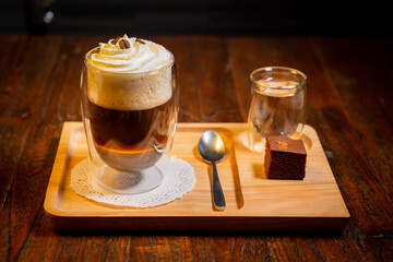 Glass of coffee topped with whipped cream and coffee beans, a glass of water, spoon and a brownie on a wooden tray