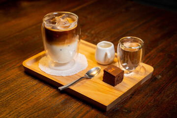 High angle shot of a glass of iced coffee with milk, ice cubes, a small syrup pitcher, a glass of water, spoon and a brownie on a wooden tray