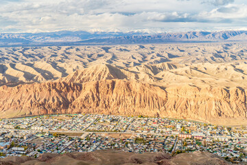 A striking aerial view of a small town nestled at the foot of rugged red cliffs and surrounded by an expansive desert-like landscape. The intricate patterns of the rolling hills and distant mountains 