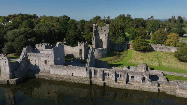 Desmond Castle, Adare, County Limerick, Ireland, September 2024. Drone orbits clockwise and rises above from River Maigue with clear reflections of grey broken walls of the 13th century Norman ruin.