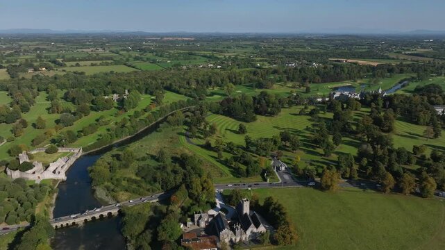 Adare, County Limerick, Ireland, September 2024. Drone orbits clockwise above the Maigue river as cars cross the old stone bridge with church and castle ruins on the scenic riverbanks.