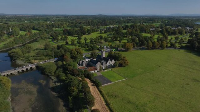 Augustinian Friary, Adare, County Limerick, Ireland, September 2024. Drone orbits clockwise at a high angle crossing the Maigue river under a blue sky showcasing building, bridge, and grassy fields.