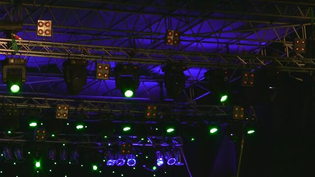 Stage lighting setup with colorful green and orange lights at a concert event highlighting the metal rigging structure and spotlights under a dark background