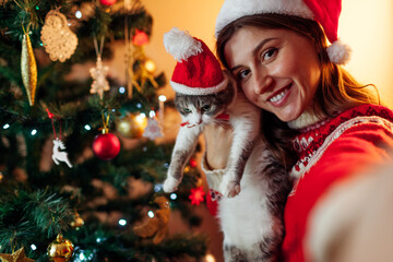 Christmas selfie with cat by New Year tree at home. Woman holding pet with Santa's hat on taking festive picture