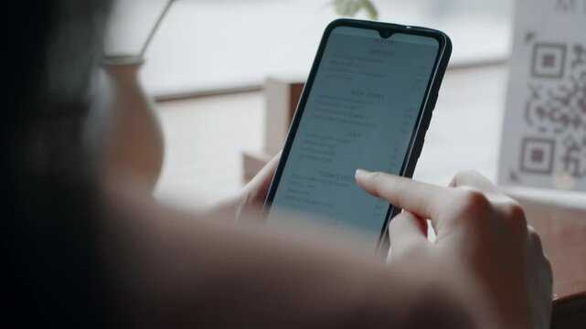 Close-up over the shoulder shot of unrecognizable woman reading menu on smartphone, while sitting in restaurant