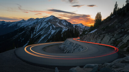 A picturesque winding mountain road at sunset, showcasing vibrant red and orange trails of car lights against snow-capped peaks.