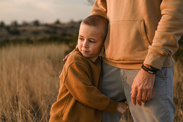 A boy gazes at the sunset while lovingly holding his father’s leg. The warm glow of the setting...