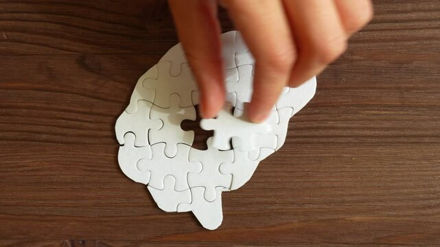 Close-up of a hand inserting a puzzle piece into a brain shape formed by other pieces on a wooden surface, symbolizing cognitive processes.