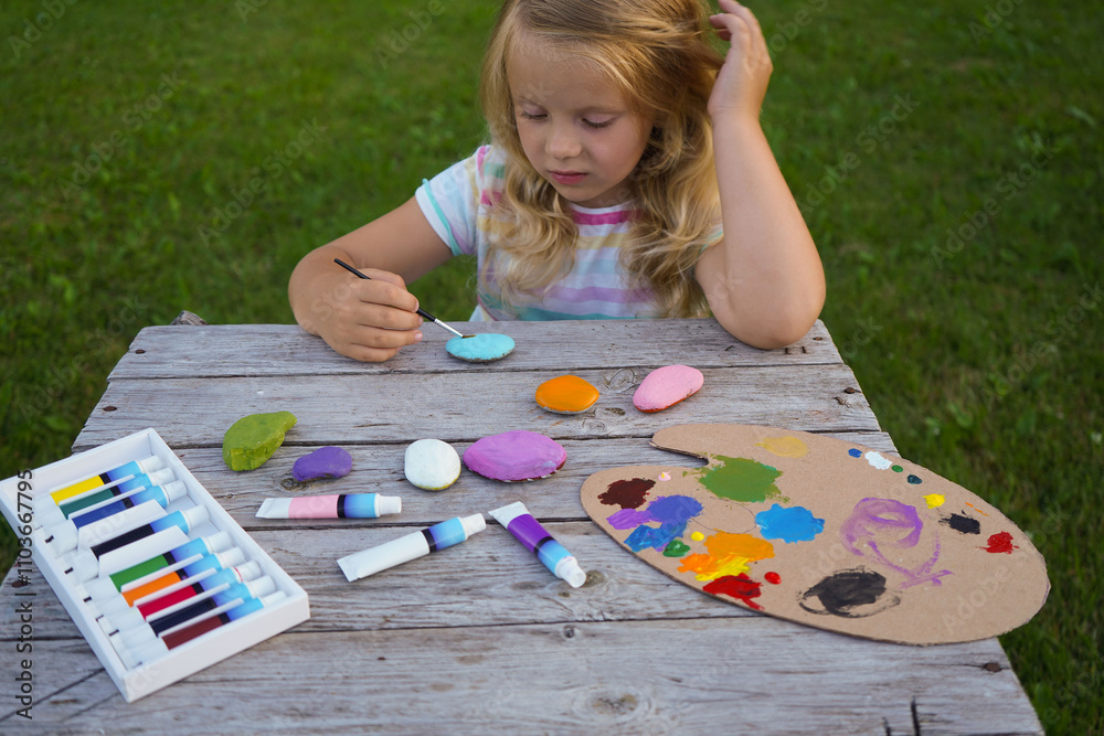 Wall mural Outdoor workshop in summer. A selective focus of a caucasian girl holding a painted flat stone in his hands. Do it yourself.