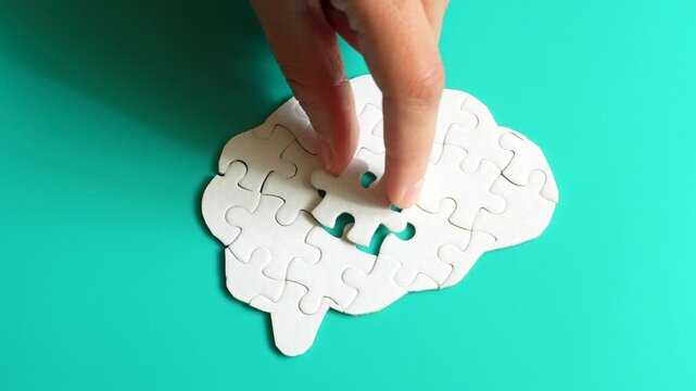 Hand placing a puzzle piece into a brain shape made of interlocking pieces on a teal background, symbolizing mental engagement.
