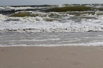 Dynamic view of ocean waves crashing onto a sandy beach, showcasing frothy white foam and clear blue water under sunlight