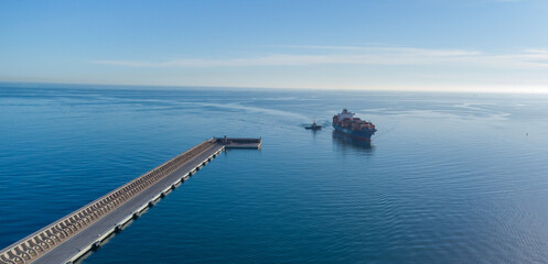Obraz premium Majestic cargo ship sails under clear skies, nearing a coastal pier against a tranquil sea backdrop?ideal for illustrating global trade, maritime industry, or serene ocean travel concepts.