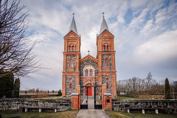 A stunning brick and stone church with twin spires, surrounded by a stone fence, under a dramatic cloudy sky in a rural setting..