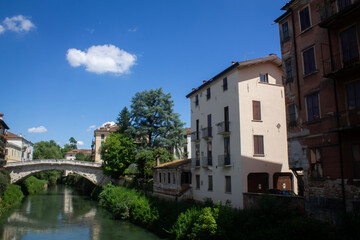 A small bridge and a river next to a street and facades of houses in the Italian city of Vicenza in the Veneto region in summer against a background of blue sky and green trees.