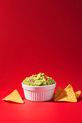 white ramekin filled with fresh guacamole sits against vibrant red background, accompanied by crispy tortilla chips, creating delightful and appetizing scene