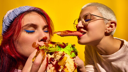 Two women emotional and passionately eating hot dog with ketchup and lettuce, against bright yellow...