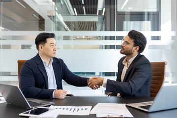 Fototapeta premium Businessmen shaking hands in modern office. Image captures partnership, trust, successful deal. Laptops, charts, documents on desk highlight professional environment. Concept of teamwork, cooperation