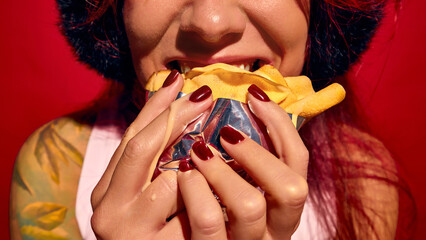 Cropped image of cheerful young woman eating crepe, pancake wrapped in foil, set against red background. Outdoor food. Concept of street food, winter fairs, pop art, holiday snacks