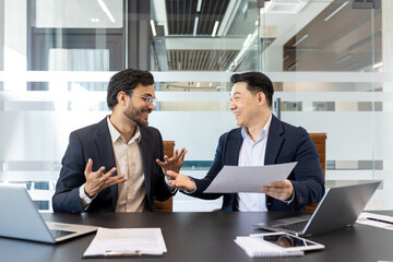 Businessmen conversing in office while reviewing documents, showcasing teamwork and collaboration. Smiling professionals fostering communication and productivity. Laptops and notes on table.