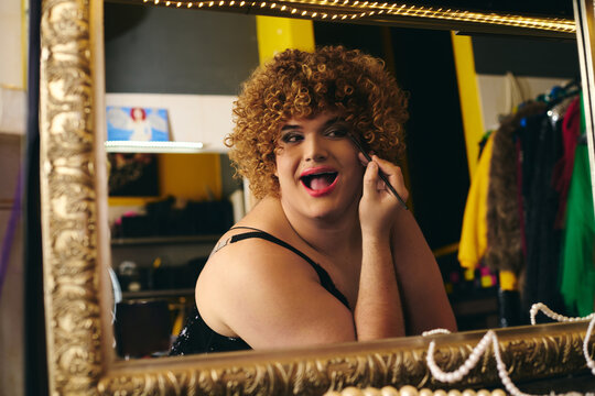 A drag queen applies makeup in a bustling backstage dressing room filled with holiday cheer.
