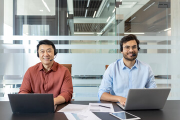 Two male customer service professionals with headsets work at laptops in stylish office. They focus on customer support, technology utilization, enhancing client satisfaction, and teamwork.