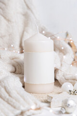 Pillar candle with label near white sweater and Christmas decor and lights, mockup