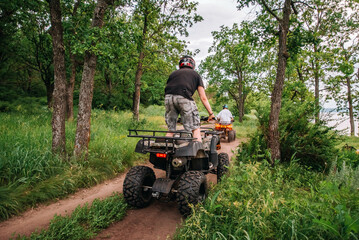 Fototapeta premium a girl and a guy bikers ride quad bikes in a beautiful green forest on a river cliff, their dogs are running nearby