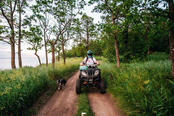 a girl and a guy bikers ride quad bikes in a beautiful green forest on a river cliff, their dogs are running nearby