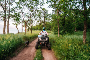 a girl and a guy bikers ride quad bikes in a beautiful green forest on a river cliff, their dogs are running nearby
