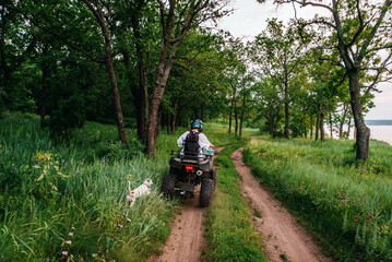 a girl and a guy bikers ride quad bikes in a beautiful green forest on a river cliff, their dogs are running nearby