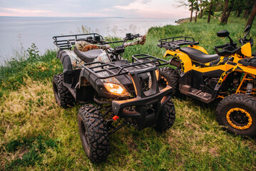 Fototapeta premium quad bikes stand on the river bank in the forest against the backdrop of a beautiful evening sky and water. headlights wheels muffler close-up