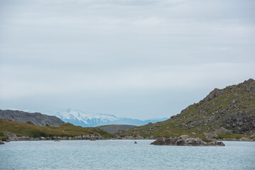 Dramatic view to rocky isle on blue mountain lake against snow covered mountain range far away in rainy weather. Ripples on azure water surface of alpine lake in high mountains under cloudy gray sky.