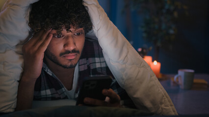 Gadget internet social media addict guy Latino tired man Arabian confused worried puzzled Indian male at night evening dark home under blanket cover with duvet scrolling mobile phone upset frustrated