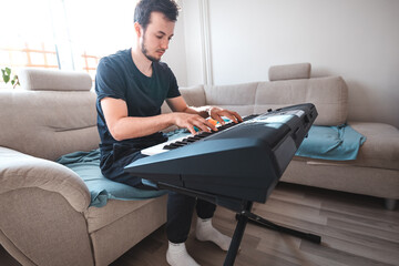 Man playing a digital keyboard in a relaxed home setting, seated on a sofa with natural light...